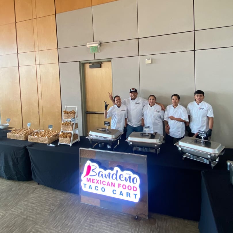 Bandeño Mexican Food staff smiling behind a taco cart with chafing dishes and food baskets.