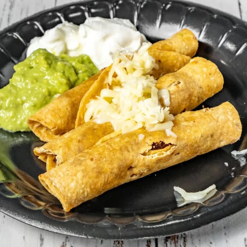 Bandeño Mexican Food taquitos with guacamole, sour cream, and shredded cheese on a black plate.