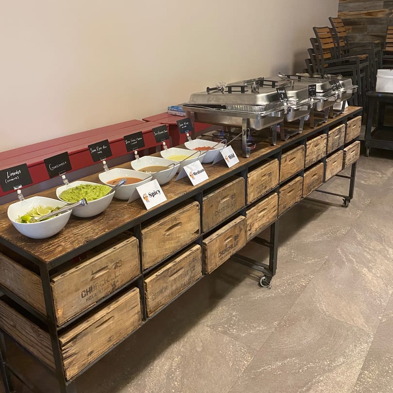 A Mexican food buffet with bowls of guacamole, salsa, and limes, alongside chafing dishes.