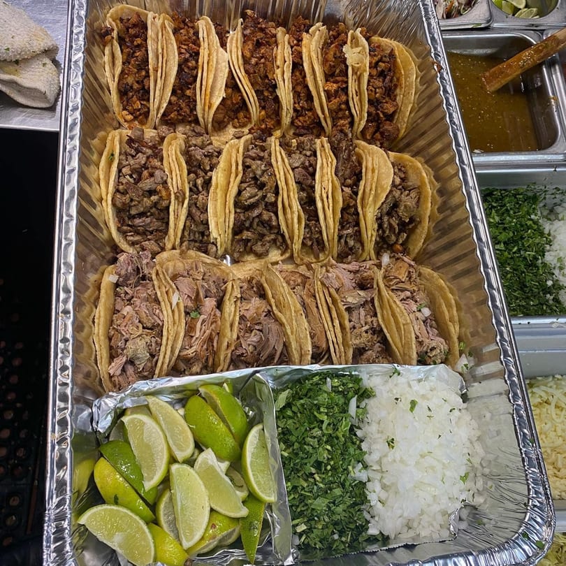 A catering tray of Bandeño Mexican Food tacos with various meats, limes, cilantro, and onions.
