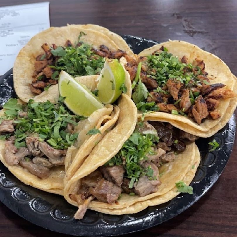 Four Bandeño Mexican Food tacos with meat, cilantro, and lime wedges on a black plate.