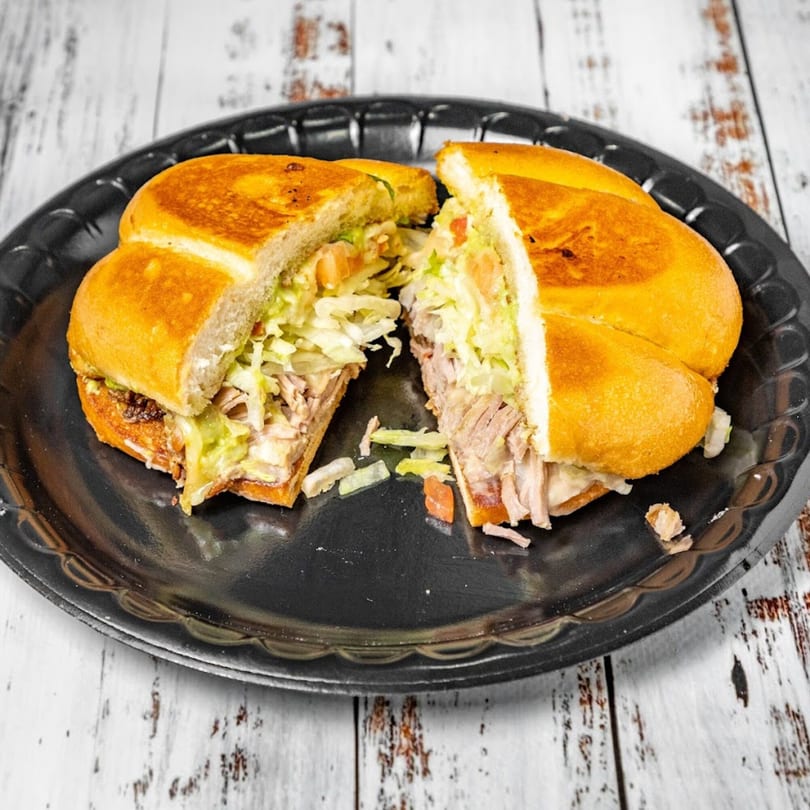 A Bandeño Mexican Food torta, cut in half, filled with meat, lettuce, and tomato on a black plate.