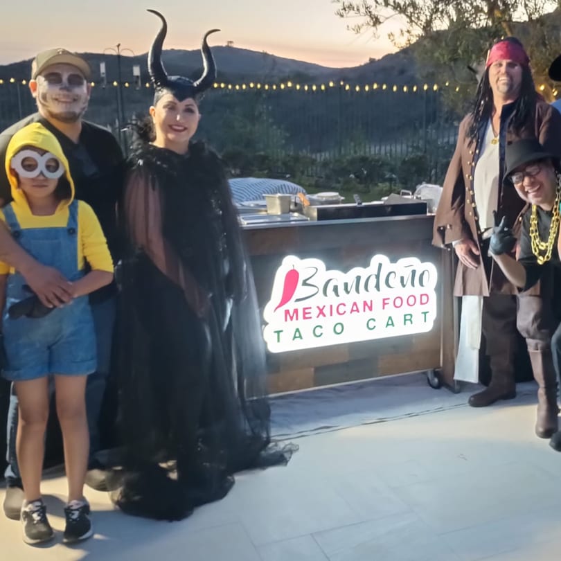 Four people in Halloween costumes stand around a Bandeño Mexican Food taco cart outdoors at dusk.