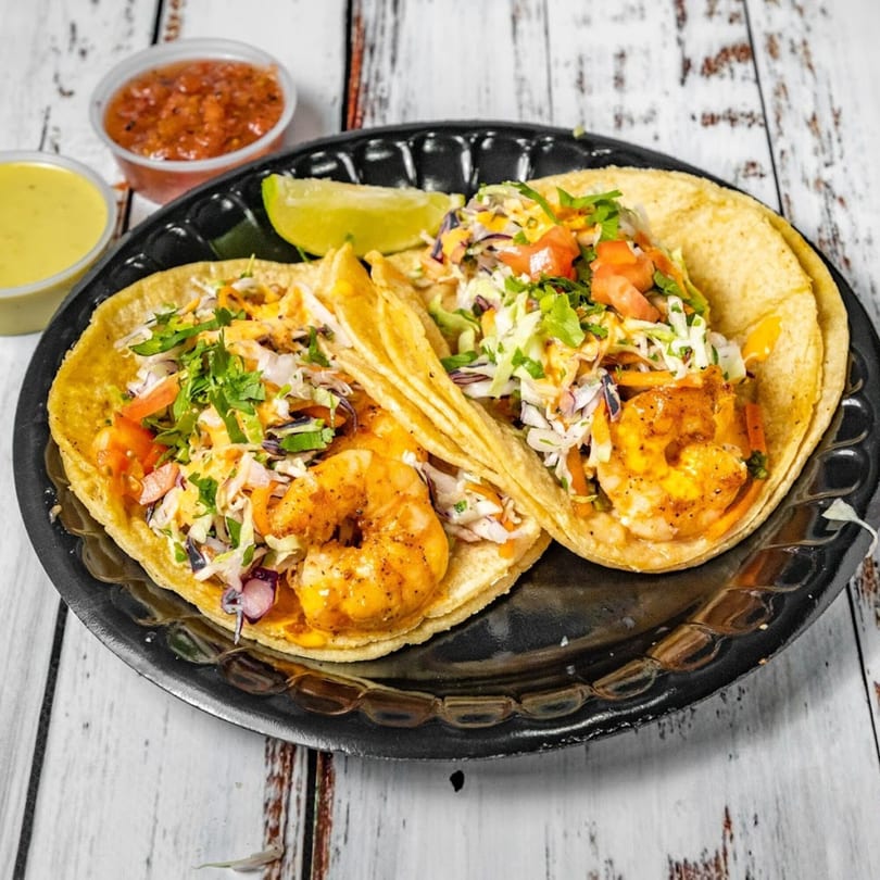 Two Bandeño Mexican Food shrimp tacos with cabbage, tomatoes, and cilantro, served with salsa and sauce.